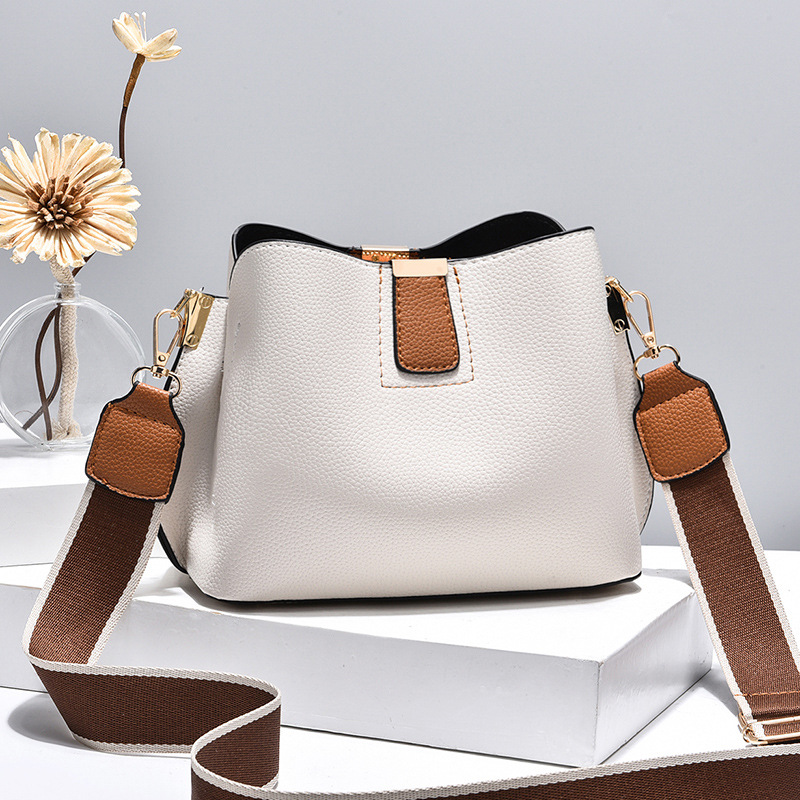 Minimalistyczna torebka typu bucket bag z kontrastowymi kolorami paska i elementami metalowymi, miękka skóra o delikatnej fakturze, regulowany pasek crossbody do codziennego noszenia.